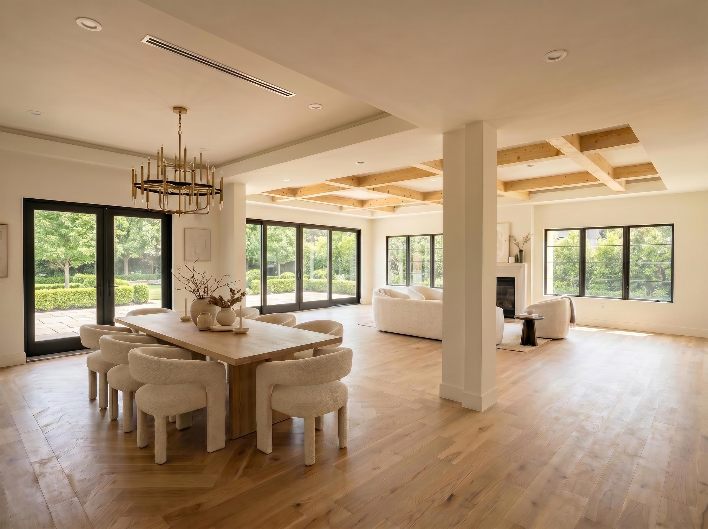 Staged living and dining room with a dining table and boucle chairs beneath the chandelier, herringbone oak floors, coffered wood ceiling, and black-framed doors opening to a finished garden.